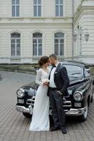 elegante linda noiva e belo noivo abraçando no elegante carro preto na luz. vista incomum de volta. casal de noivos de luxo em estilo retrô. foto