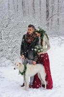 linda noiva e noivo com um cachorro branco estão de pé no fundo de uma floresta de neve. foto