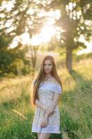 retrato de uma jovem linda em um vestido de verão. sessão de fotos de verão no parque ao pôr do sol. uma garota se senta debaixo de uma árvore na sombra.