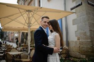 um casal sorridente está desfrutando de bebidas em uma mesa de bar ao ar livre na cidade. o homem está sentado à mesa olhando para um tablet digital e a mulher está de pé perto dele. foto