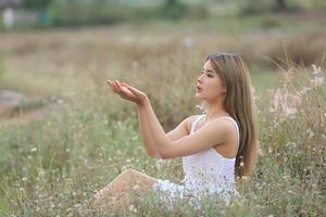 bela jovem sentada no campo na grama verde e soprando dente de leão. ao ar livre. Aproveite a natureza. menina sorridente saudável no gramado da primavera. conceito livre de alergia. liberdade foto