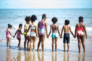 crianças brincando correndo na areia na praia, um grupo de crianças de mãos dadas na praia no verão, vista traseira contra o mar e o céu azul foto