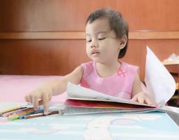 Menina asiática de 3 anos de idade escolhendo lápis coloridos para pintar no livro de colorir em casa. foto