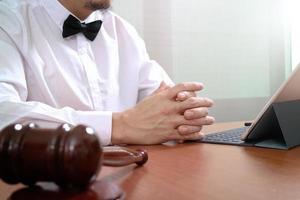 contexto de justiça e direito. mão de advogado masculino trabalhando com telefone inteligente, teclado de encaixe de computador tablet digital com martelo e documento na mesa de madeira foto