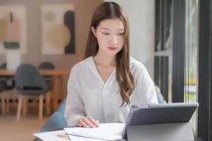 mulher confiante asiática veste uma camisa branca está usando um tablet para trabalhar e há muitos documentos em cima da mesa. seu rosto sorrindo em uma sala de trabalho em casa.