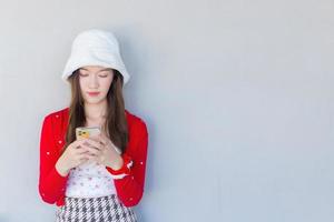 linda mulher asiática sorrindo que usa casaco vermelho e chapéu branco como uma garota santa segura e joga um smartphone na mão sobre o fundo branco. foto