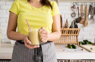 jovem morena bebendo smoothie de banana de frutas na cozinha de casa foto