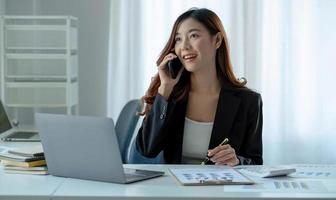 mulher de negócios asiáticos tem a alegria de falar ao telefone, laptop e tablet na mesa do escritório.