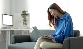 jovem mulher asiática freelance de negócios trabalhando no laptop verificando as mídias sociais enquanto estava deitado no sofá quando relaxa na sala de estar em casa. foto