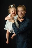 um pai segura nos braços sua filhinha de um ano. filmado em um fundo preto.
