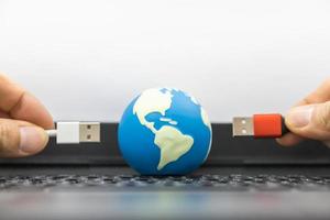 comunicação, computador e conceito de tecnologia global. close-up da mini bola mundial com a mão segurando o cabo usb no teclado do computador portátil e copie o espaço. foto