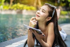 linda mulher asiática de bom humor jogando um tablet à beira da piscina e pense no passado que o fez feliz foto