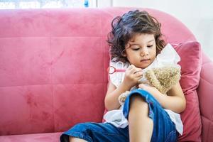 uma garotinha bonita gosta de jogar brinquedo de seringas de injeção de brinquedo no ursinho de pelúcia no sofá em casa, tempo de educação em casa foto