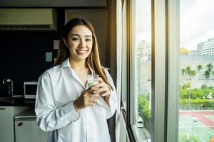 jovem mulher asiática, olhando pela janela, aproveitando o novo dia, sentindo-se descansado e bebendo um copo de leite no hotel. foto