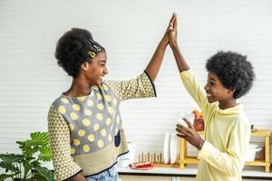 crianças bonitinhas tomando café da manhã com leite na cozinha, menino bonito afro-americano bebendo leite com sua irmã, amizade adolescente menino e menina estão batendo as palmas das mãos, conceito de comida e bebida foto