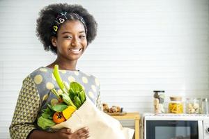 feliz garota afro-americana saudável na cozinha, adorável garota sorrindo e segurando o saco de papel com alimentos frescos cheios de legumes, alimentação saudável foto