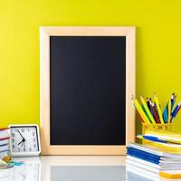 quadro-negro, livros didáticos, relógio e material escolar na mesa pela parede amarela. vista lateral, espaço para texto. de volta ao conceito de escola. foto