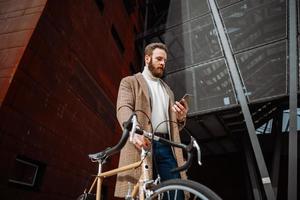 jovem com bicicleta segurando um smartphone na frente do prédio. empresário criativo em uma área de negócios moderna. foto