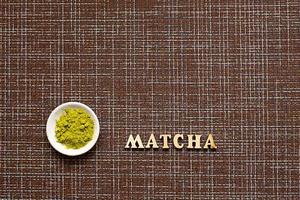 chá matcha em pó verde japonês em uma travessa, sobre um fundo marrom. inscrição das letras de madeira em inglês. pó solto. espaço para texto. cozinhar, um ingrediente para uma bebida revigorante foto