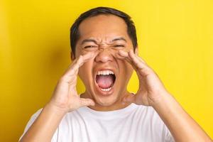 retrato close-up de homem asiático com gesto de grito, xícara de mãos ao redor da boca isolada em fundo amarelo foto