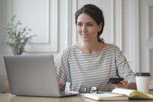 jovem italiana freelancer segurando cartão de crédito, usando laptop ao fazer pagamentos online foto