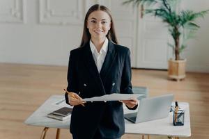 jovem mulher de negócios sorridente olhando para a câmera e segurando o documento na frente da mesa de escritório foto