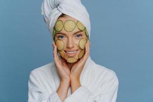 jovem e bela mulher aplica fatias de pepino no rosto tem pele limpa perfeita toca bochechas suavemente vestida com roupão confortável tem toalha enrolada na cabeça. conceito de cosmetologia e spa foto