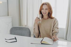 mulher ruiva feliz com copo de água mineral pura, escrevendo a lista de compras no caderno em casa foto