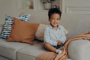 menino afro doce em roupas casuais, sentado no sofá confortável e jogando dentro de casa foto