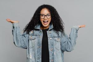 retrato de mulher afro-americana encaracolada satisfeita feliz levanta as palmas das mãos, finge segurando algo, usa óculos ópticos e jaqueta jeans, isolada em fundo cinza. conceito de pessoas e felicidade foto