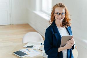 próspera líder feminina da equipe de trabalho detém dispositivo tablet digital, desenvolve ideias de negócios, tem sorriso cheio de dentes, cabelo ruivo, veste roupas elegantes, fica em seu próprio gabinete, envolvido no processo de trabalho foto