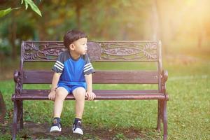 menino tailandês sente-se no banco no parque com fundo verde de botânica foto