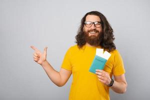 homem jovem hippie com cabelo comprido, segurando o passaporte com bilhetes apontando para copyspace.