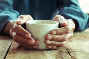 mãos de velho segurando a xícara de café na mesa de madeira. foto