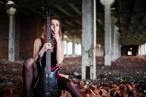 garota punk de cabelo vermelho usa saia preta e vermelha, com baixo em lugar abandonado. retrato do cigarro de músico de mulher gótica. foto