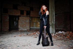 garota punk de cabelo vermelho usa preto com guitarra baixo em lugar abandonado. retrato de músico de mulher gótica. foto