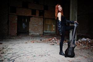 garota punk de cabelo vermelho usa preto com guitarra baixo em lugar abandonado. retrato de músico de mulher gótica. foto