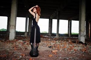 garota punk de cabelo vermelho usa preto com guitarra baixo em lugar abandonado. retrato de músico de mulher gótica. foto