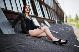 garota modelo sexy com pernas longas em roupa de lingerie preta corpo maiô combidress e jaqueta posada no telhado do local industrial abandonado com janelas. foto