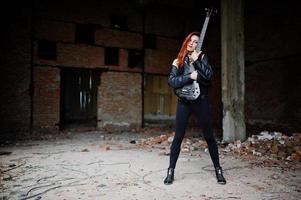 garota punk de cabelo vermelho usa preto com guitarra baixo em lugar abandonado. retrato de músico de mulher gótica. foto