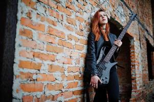 garota punk de cabelo vermelho usa preto com guitarra baixo em lugar abandonado. retrato de músico de mulher gótica. foto