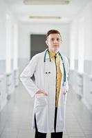 bonito e confiante jovem médico de jaleco branco com estetoscópio posando no hospital. foto