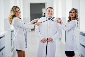 duas belas médicas e um homem de jaleco branco posando no hospital com estetoscópio. foto