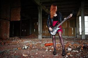 garota punk de cabelo vermelho usa saia preta e vermelha, com baixo em lugar abandonado. retrato de músico de mulher gótica. foto