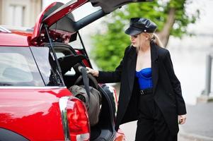 retrato do modelo de mulher loira bonita moda sexy no boné e todo preto com maquiagem brilhante perto do carro vermelho da cidade colocar a bolsa no porta-malas. foto