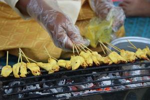 espetadas de frango estão grelhando no torrador tradicional com a mão pegando a vara de espetadas e fogo de carvão, tailândia. foto