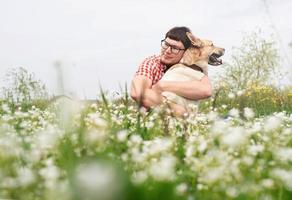 homem feliz sentado com cão pastor de raça mista na grama verde em flores da primavera foto