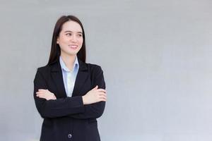 mulher asiática de trabalho de negócios profissional que usa um terno formal preto fica com o braço cruzado sorri alegremente no escritório como pano de fundo. foto