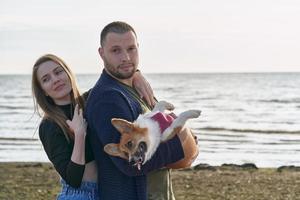 jovem casal feliz com cachorro dançando na praia. linda garota e cara e cachorro corgi se divertindo foto