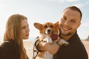 jovem casal feliz com cachorro tomando selfie na praia. linda mulher e homem e corgi foto